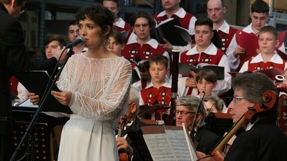 Schauspielerin Friederike Becht steht in weißem Kleid vor einem Orchester.