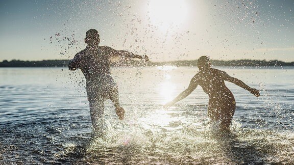 ein Mann und eine Frau laufen bei Sonnenuntergang ins spritzende Wasser