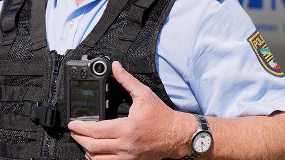 Ein Polizist bedient symbolisch eine Bodycam, die an seiner Schutzweste befestigt ist.
