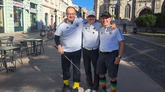 Drei Männer in weißen T-Shirts stehen mit Golfschläger in einer Innenstadt.