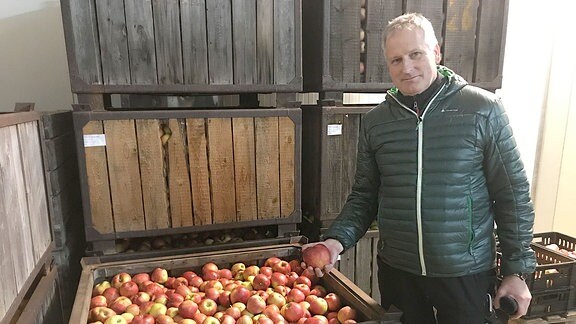 Mann steht neben einer großen Kiste mit Äpfeln und hält einen Apfel in der Hand.