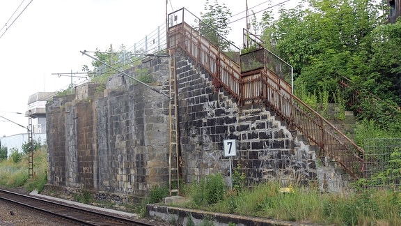 Entlang einer elektrifizierten Bahnstrecke führt eine Treppe mit rostigem Geländer eine Mauer hinauf.