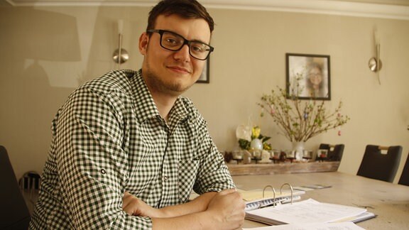 Ein junger Mann mit kariertem Hemd sitzt an einem Tisch vor Unterlagen und blickt in die Kamera.