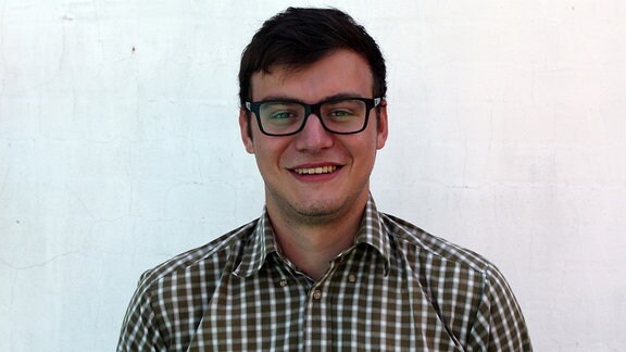 Porträtaufnahme eines jungen Mannes, dunkle Haare, schwarze Brille und kariertes Hemd, der vor einer weißen Wand steht und lächelt