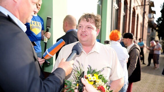 Hannes Loth in weißem Hemd mit Blumenstrauß in der Hand spricht in ein MDR-Mikrofon.