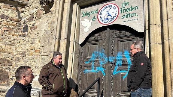 Drei Männer stehen vor einer mit FCM-Schriftzug besprühten Kirchentür.