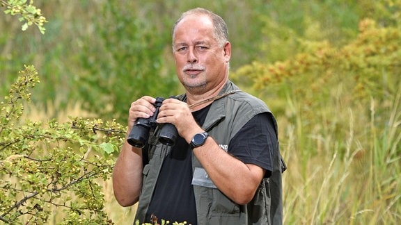 Ein Mann in schwarzem T-Shirt und schwarzer Weste steht mitten in der Natur. Er schaut in die Kamera, hat ein Fernglas in den Händen.