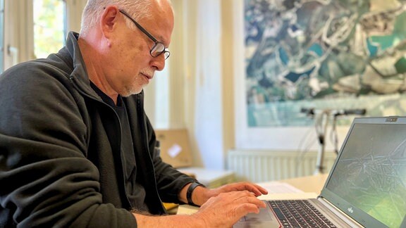 Ein Mann mit Brille, bekleidet mit in einer dunkles Fleece-Jacke sitzt vor einem Laptop und arbeitet
