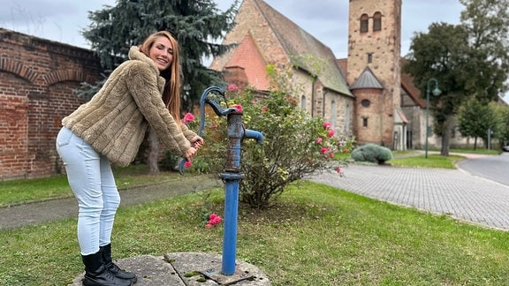 Eine Frau steht vor Backsteingebäuden an einer alten Wasserpumpe