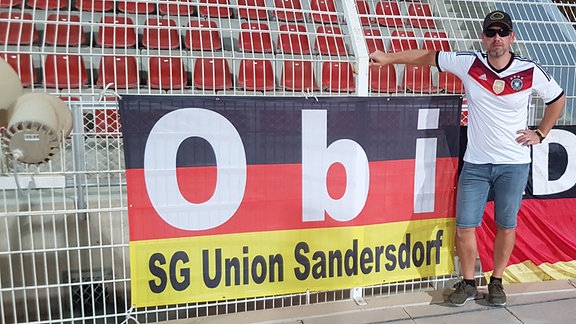 Ein Mann steht in einem leeren Fussballstadion vor einem Zaun an dem eine Deutschlandflagge hängt. Darauf steht sein Spitznamen "Obi".