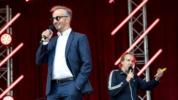Zwei Männer stehen mit Mikrofonen in der Hand auf einer Bühne