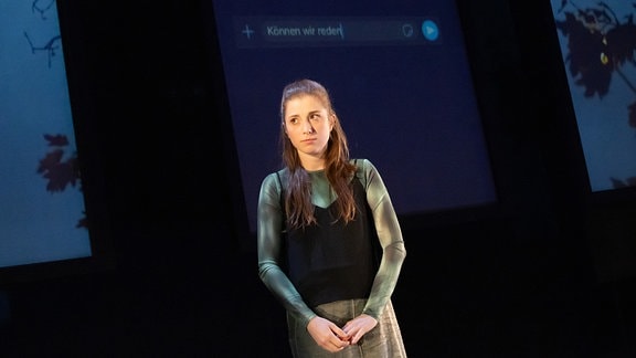Eine junge Frau mit langem braunem Haar steht auf einer Theaterbühne, im Hintergrund ist die unabgeschickte Chatnachricht "Können wir reden" auf eine Leinwand projeziert.