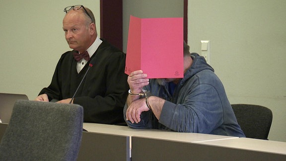 Ein Mann auf der Anklagebank verdeckt sein Gesicht mit einem roten Ordner
