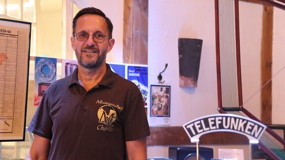 Ein Mann mit Brille und T-Shirt mit der Aufschrift "Museumshof Chörau" steht in einem Raum mit alten Radios