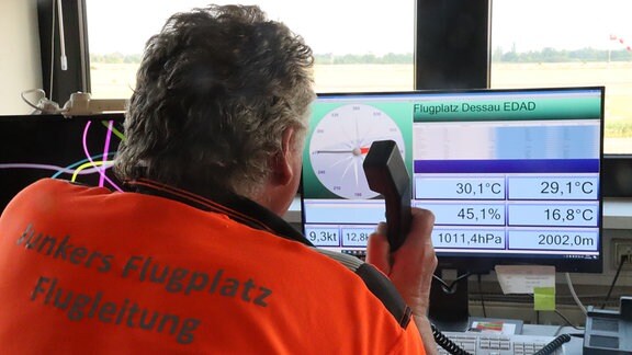 Ein Mann beobachtet Wetterdaten auf einem Bildschirm, auf seinem Rücken steht "Junkers Flugplatz Flugleitung"