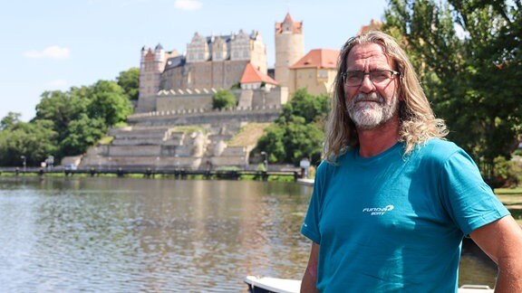 Ein Mann mit blauem T-Shirt und schulterlangem Haar steht an der Saale, im Hintergrund erwächst das Schloss Bernburg