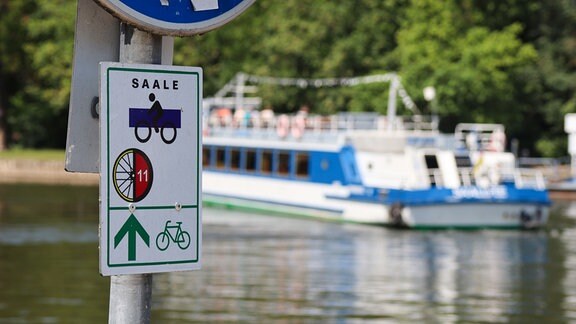 Ein Schild mit Fahrradsymbolen und der Aufschrift "Saale" steht am Fluss, auf dem ein Schiff fährt