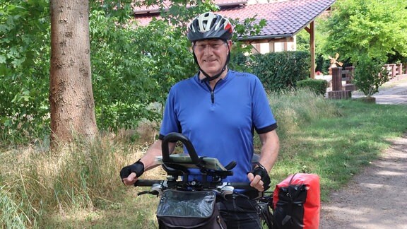 Ein Mann mit Helm und blauem Shirt hält sein Fahrrad und lächelt in die Kamera