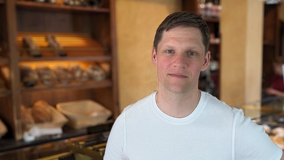 Ein Mann mit weißem T-Shirt steht vor einem Regal mit Broten