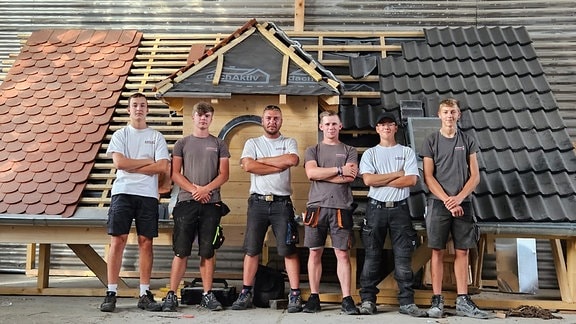 echs Handwerker-Auszubildende einer Dachdeckerei stehen in ihrer Arbeitskleidung vor dem Modell eines teilweise gedeckten Daches.