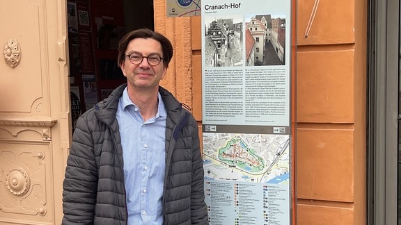 Ein Mann mit runder Brille steht vor einem Gebäude und lächelt in die Kamera.