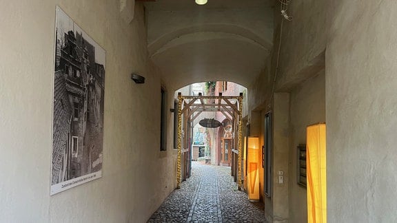 Man blickt durch die Gasse zwischen zwei grauen Häusern. Links und rechts an den Wänden hängen Gemälde