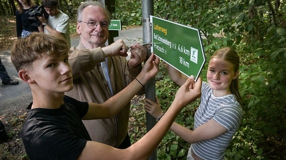 Rainer Haseloff (CDU), Ministerpräident von Sachsen-Anhalt, Jaron Held und Sophie Rommel vom Evangelischen Schulzentrum Bad Düben bringen ein neues Lutherwegschild an.