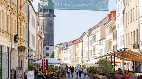 In einer Fußgängerzone in Lutherstadt Wittenberg laufen mehrere Menschen, andere sitzen in Cafés am Straßenrand.