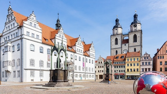Auf einem Marktplatz sind neben einem Brunnen und einer Skulptur auch Rathaus und Türme einer Kirche zu sehen.