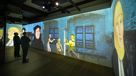 Projektionen von Zeichnungen Luthers und seiner Frau sowie eines historischen Raumes sind in einer Ausstellung zu sehen, Besucher der Ausstellung betrachten die Installation.