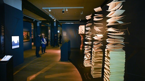 Blick in die Ausstellung "Buchstäblich Luther" mit schwarzen Wänden und einzelnen Exponaten. Besucher im Hintergrund betrachten eine Ausstellungswand. Im Vordergrund sind Blätter mit einer Schnur verbunden gestapelt.