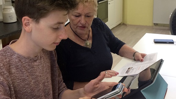 Jugendlicher mit Handy in der Hand sitzt neben älterer Frau vor einem Tablet.