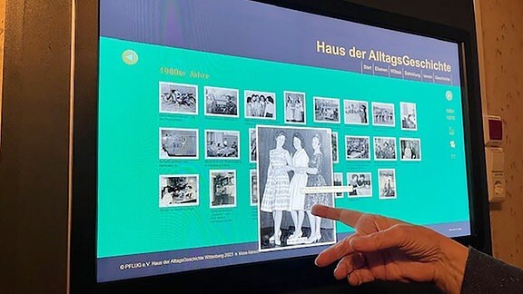 Eine Hand zeigt auf ein Tablet in einer Ausstellung, auf dem mehrere Fotos abgebildet sind.