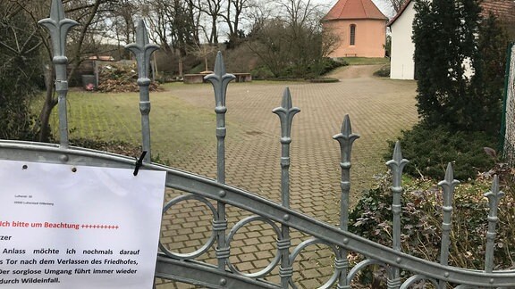 Provisorisches Schild an Zaun zu Friedhof mit Kapelle.