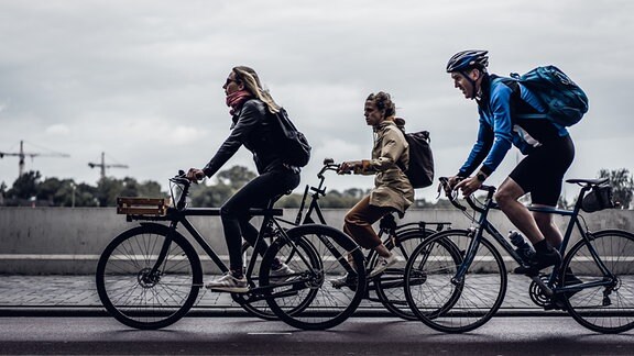 Zwei Frauen und ein Mann fahren mit dem Fahrrad auf dem Radweg durch eine Stadt.