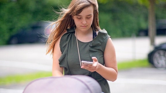 Junge Frau schiebt einen Kinderwagen und schaut auf ihr Handy.