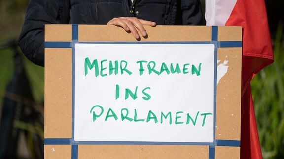 Teilnehmerin einer Kundgebung steht hinter einem Plakat mit der Aufschrift „Mehr Frauen ins Parlament“