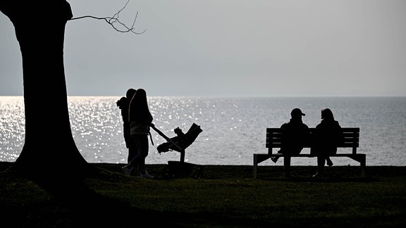 Ein Mann und eine Frau sitzen am späten Nachmittag am Ufer eines Sees auf einer Bank. Daneben gehen Menschen mit einem Kinderwagen.