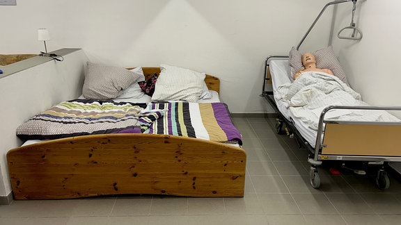 In einem steril eingerichteten Zimmer mit weißen Wänden stehen ein normales Bett und ein Krankenbett, in dem eine Puppe liegt. 