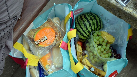 In zwei Einkaufstaschen sind mehrere Lebensmittel verstaut, darunter Weintrauben, Bananen und eine Melone