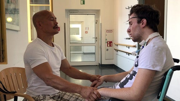 Zwei Männer machen Atemübungen in einem Krankenhausraum. Der Mann rechts sitzt in einem Rollstuhl und trägt ein Atemgerät