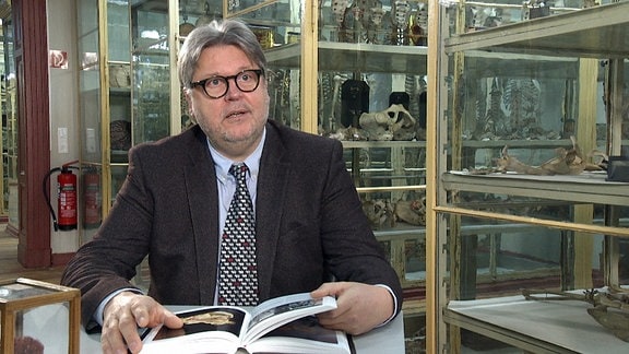 Ein Mann mit Anzug sitzt in einem Anatomie-Raum und blättert durch ein Buch
