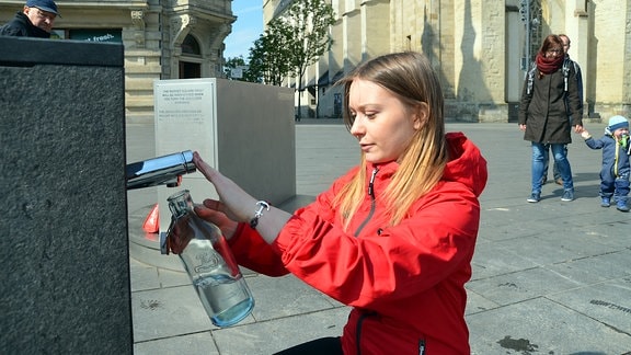 Eine junge Frau füllt Wasser aus einem Trinkwasserbrunnen in eine Glasflasche ab