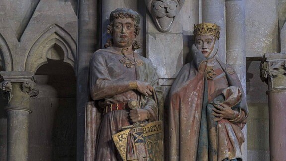Ganzkörperskulpturen von einer Mann und einer Frau mit Kronen und bunten Gewändern an einer Kirchenwand.