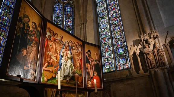 Ein dreiteliges Gemälde in einem Dom, dahinter große Kirchenfenster und rechts an der Wand Stifterfiguren aus Stein
