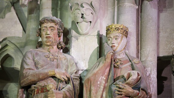 Zwei Figuren aus Stein, ein Mann und eine Frau mit goldener Krone und bis über die Wangen gezogenem Umhang