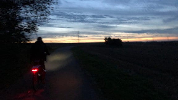Abendstimmung: Ein Fahrradfahrer fährt mit Rücklicht in der Dunkelheit, am Horizont sieht man die untergehende Sonne und am Himmel noch einige Wolken