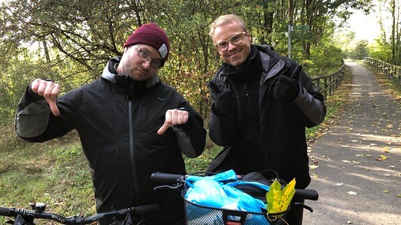 Leonard Schubert und André Plaul mit Fahrrädern auf einem Radweg. Leonard gestikuliert "Daumen runter", André "Daumen rauf"