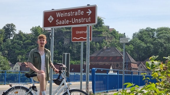 Paul Lüder von hinreisend auf einem Fahrrad vor einem Schild mit der Aufschrift "Weinstraße Saale-Unstrut", das auf einer Brücke steht