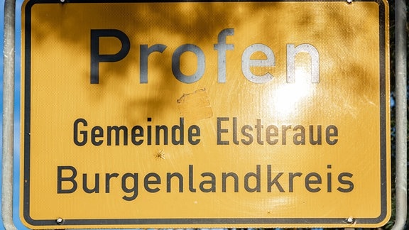 Nahaufnahme eines gelben Ortsschildes, auf dem in schwarzer Schrift -Profen, Gemeinde Elsteraue, Burgenlandkreis, geschrieben steht.